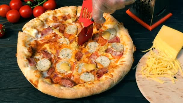 Die Hände schneiden unbeholfen Pizza ab, auf Scheiben, mit Wurst, Tomate und Käse, mit speziellem Messer - Schneidrad. Auf dem Holztisch liegen auch Tomaten und geriebener Käse. - Filmmaterial, Video