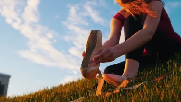 Young attractive ballerina takes on pointe shoes and wears a sitting on the green hill. - Footage, Video