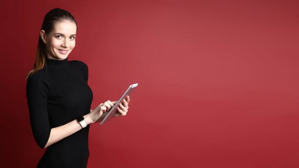 Портрет красивой улыбающейся кавказки в черном платье с помощью планшета на красном фоне
 - Фото, изображение