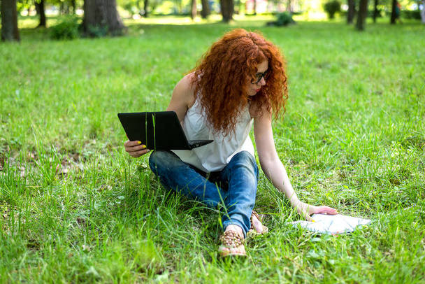 Woman sitting on grass at park working on laptop. Red-haired female freelancer working outdoors - Fotó, kép