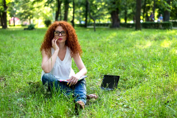 Kobieta siedzi na trawie w parku pracy na laptopie. Rudowłosy kobieta Freelancer pracy na zewnątrz - Zdjęcie, obraz