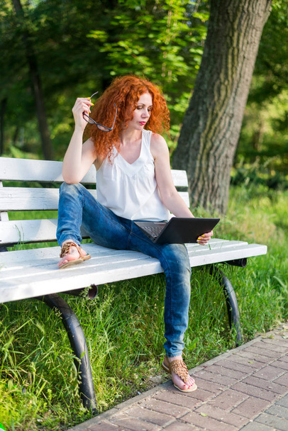 Red-haired female freelancer working on a laptop while sitting on a park bench. - Zdjęcie, obraz