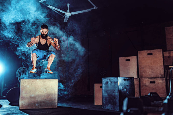 Fit tattoed bearded man jumping onto a box as part of exercise routine. Man doing box jump in the gym. Athlete is performing box jumps - 写真・画像