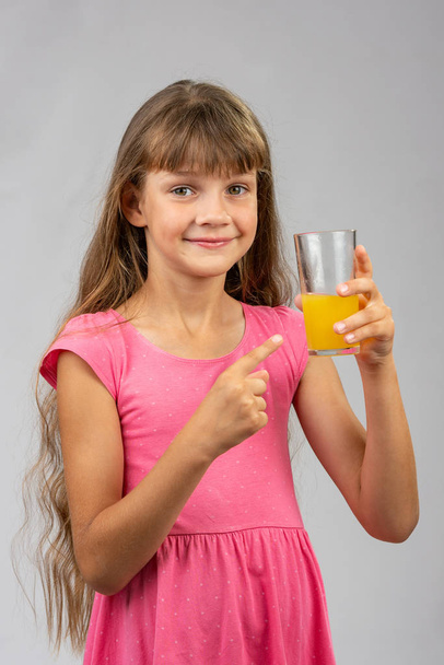 A girl holds a glass of orange juice in her hand and points her finger at it - Photo, Image