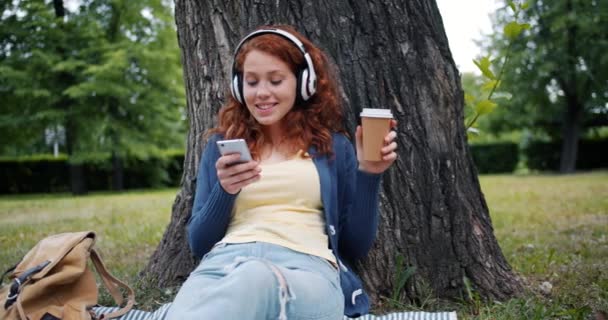 Smiling girl listening to music in headphones using smartphone relaxing in park - Metraje, vídeo