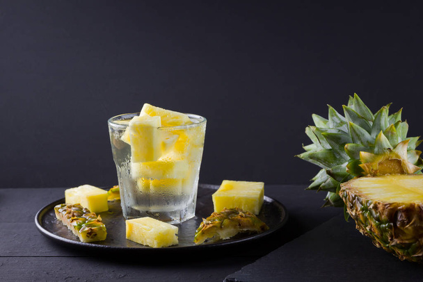 Pineapple on a dark background. Sliced pineapple on a black plate. Drink with ice and pineapple slices. Infused water. Copy space - 写真・画像