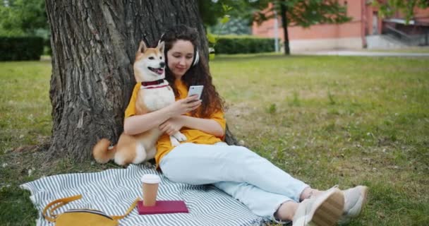 Modern girl listening to music with headphones using smartphone in park with dog - Filmati, video