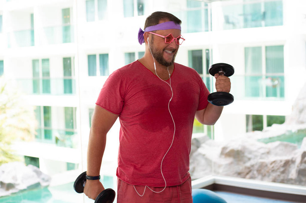 Engraçado macho gordo em óculos rosa e em uma camiseta rosa envolvida com halteres no ginásio retratando uma garota
 - Foto, Imagem
