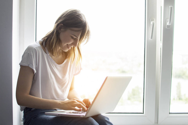young girl sitting near the window with a laptop, she uses the computer at home on the windowsill, a woman freelance, copy space for text - Photo, image