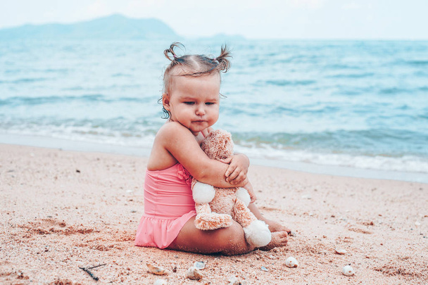 pensive little girl hugging teddy bear and looking away while sitting on seashore. - Photo, Image