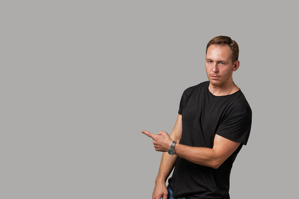 Een man in een zwart T-shirt toont met zijn handen op uw tekst of uw advertentie op een grijze achtergrond. Concept banner voor reclame, kopieer ruimte, grijze achtergrond, commercieel ontwerp. - Foto, afbeelding