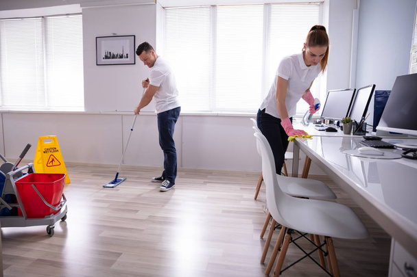 Two Smiling Young Janitor Cleaning The Desk And Mopping Floor In The Office - 写真・画像