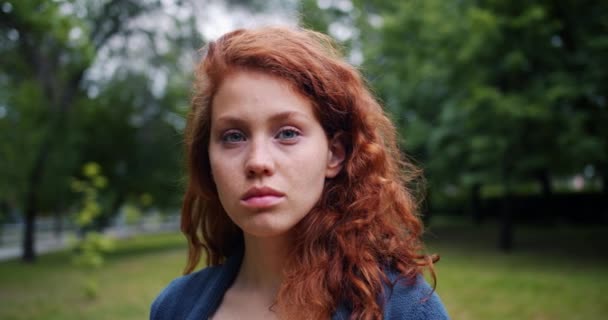Slow motion portrait of pretty serious lady with red hair standing in the park - Filmagem, Vídeo