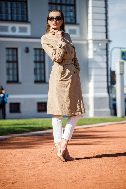 Beauty blond woman standing outdoors at daytime. Portrait of stylish model wearing beige croak, white pants and sunglasses  - Фото, зображення