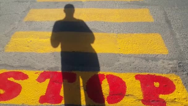 a person stands at the beginning of a pedestrian crossing, where it is written stop and waits for the passage time, on the yellow lines drawn on the asphalt, the shadow of a person is clearly visible, copy space for text - Footage, Video