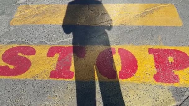 een persoon staat aan het begin van een voetgangers kruising, waar het is geschreven stop en wacht op de passage tijd, op de gele lijnen getekend op het asfalt, de schaduw van een persoon is duidelijk zichtbaar, kopieer ruimte voor tekst - Video