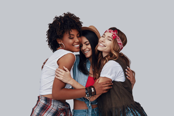 Three attractive stylish young women embracing and laughing while standing against grey background - Foto, Bild
