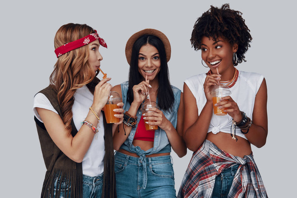 Three attractive young women drinking refreshing cocktails and smiling while standing against grey background - Valokuva, kuva