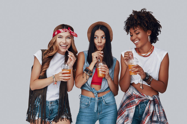 Three attractive young women drinking refreshing cocktails and smiling while standing against grey background - Foto, Imagem