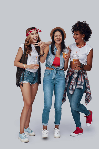 Full length of three attractive young women drinking refreshing cocktails and smiling while standing against grey background - Photo, image
