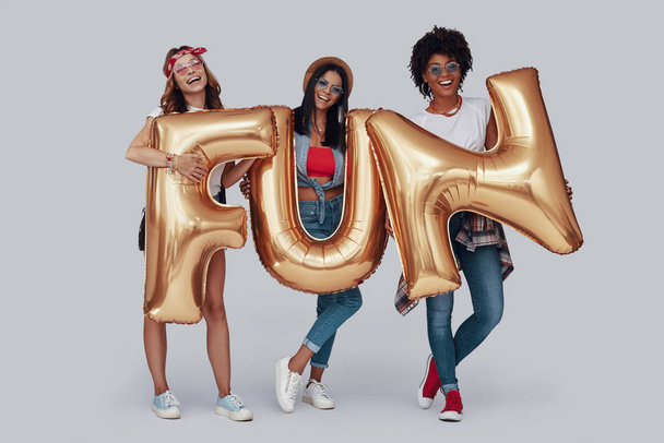 Full length of three attractive young women carrying balloons and smiling while standing against grey background - Fotó, kép