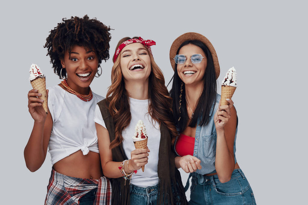 Three attractive young women eating ice cream and smiling while standing against grey background - Fotó, kép