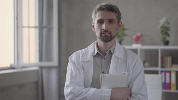 Portrait of handsome skill smiling man in white robe looking in the camera holding tablet. Adult doctor in modern private clinic. Concept of medical treatment - Кадры, видео