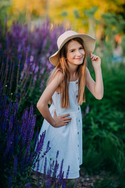 Happily smiling little girl in white dress and hat stay in the park between lavender flowers - Foto, immagini