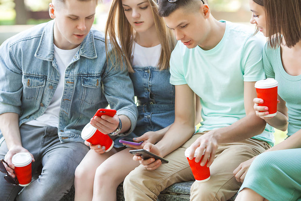 Veselí přátelé píšou do mobilu. Mladí lidé se dívají na smartphone. Skupina přátel venku s mobilními telefony a kávou. - Fotografie, Obrázek