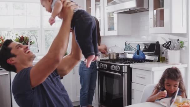 Hispanic family talking in their kitchen, mum cooking at hob, dad lifting baby in the air, close up - Materiał filmowy, wideo