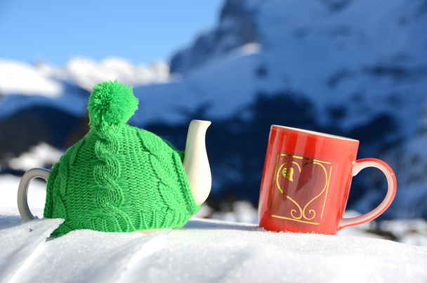 Panela de chá na tampa de malha e xícara vermelha com um coração na neve
 - Foto, Imagem