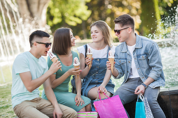 Een groep jonge vrolijke mensen die ijs eten en plezier hebben. Winkelaars buiten. Mensen na het winkelen. Grappige groep vrienden rustend bij de fontein. - Foto, afbeelding