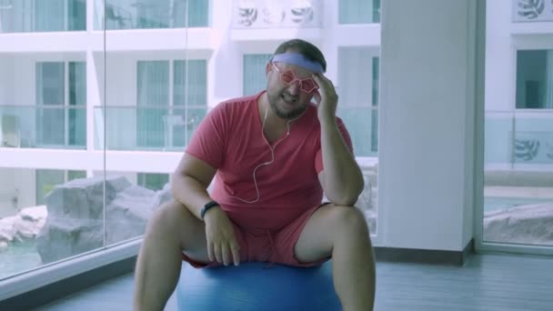 Αστείο λίπος αρσενικό σε ροζ γυαλιά και σε ένα ροζ t-shirt ασχολούνται με μια μπάλα Fit στο γυμναστήριο που απεικονίζει ένα κορίτσι. Ένας άνθρωπος φρικιό έχει πονοκέφαλο στην μπάλα στο γυμναστήριο.. - Πλάνα, βίντεο