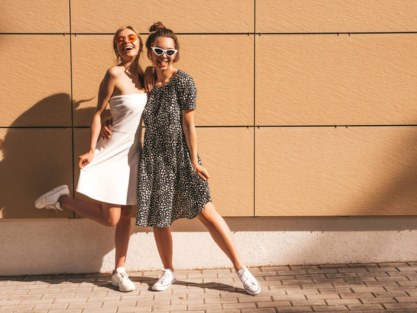 Two young beautiful smiling hipster girls in trendy summer dresses. Sexy carefree women posing in the street near yellow wall.Positive models having fun in sunglasses - Фото, изображение