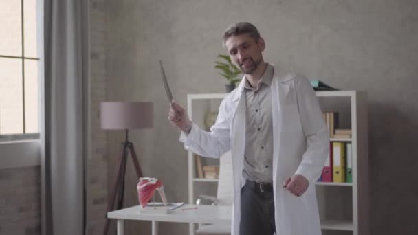 Handsome positive, successful and satisfied man in white robe dancing with x-ray in hands in modern clinic. The doctor got great news, he is happy. Concept of medicine, health care, hospital. - Metraje, vídeo