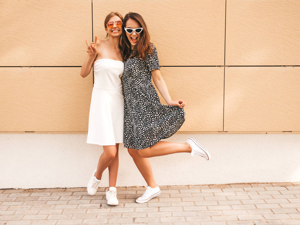 Two young beautiful smiling hipster girls in trendy summer dresses. Sexy carefree women posing in the street near yellow wall.Positive models having fun in sunglasses.Shows peace sign - Fotografie, Obrázek