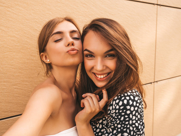 zwei junge lächelnde Hipster-Frauen in Sommerkleidung. Mädchen machen Selfie Selbstporträt Fotos auf smartphone.models posiert auf der Straße Hintergrund in der Nähe der gelben Wand.female zeigt positive Gesichtsbewegungen - Foto, Bild
