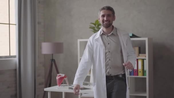 Handsome positive, successful and satisfied man in white robe dancing in the modern clinic office. Happy doctor got great news. Concept of medicine, health care, hospital. - Filmagem, Vídeo