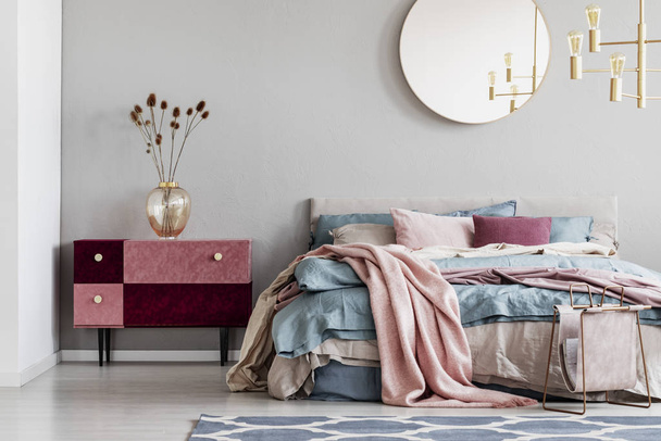 Classy round mirror on grey wall in stylish bedroom interior with warm bed with blue, pastel pink and beige bedding and velvet covered nightstand - Photo, Image