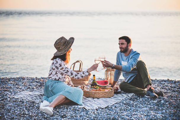 Summer beach picnic at sunset. Young happy couple having weekend picnic outdoors at seaside with bottle of sparkling wine, fruit and tray of tasty appetizers, drinking wine and looking to each other - Fotó, kép