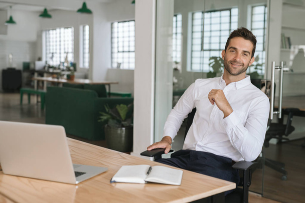 Portrait of a smiling young businessman sitting in a chair at his desk in a large modern office - Foto, imagen