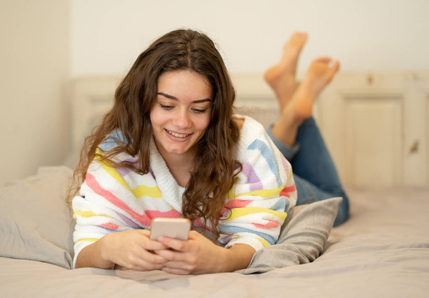 Happy pretty girl sending messages on mobile phone chatting in social media applications or gaming online lying on bed at home in people, lifestyle, communication and new technology concept. - Photo, Image