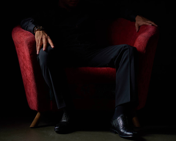 male businessman in a black suit sitting in red chair, black background, no faces visible, studio shooting - Photo, image