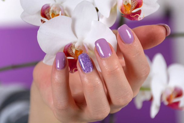 Female hand with a lilac color nails polish gel and beautiful orchid flower decoration on purple background in the studio. Manicure and beauty concept. Close up, selective focus - Photo, Image