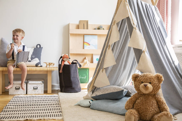 Smiling kid sitting on bench with notebook, real photo of natural playroom interior with scandinavian tent and teddy bear - Fotoğraf, Görsel