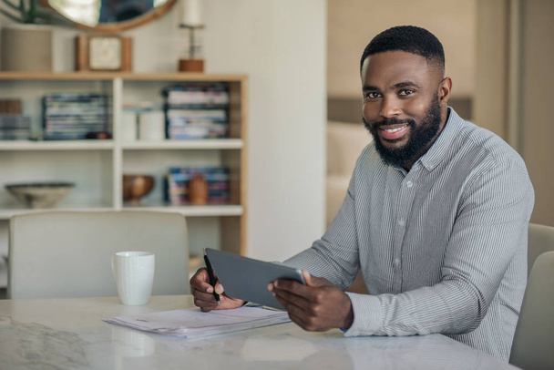 Retrato de um jovem afro-americano sorridente sentado em uma mesa em casa usando um tablet digital e analisando suas finanças
 - Foto, Imagem
