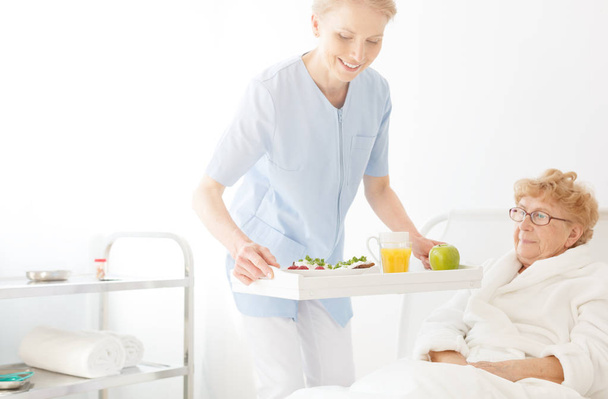 Positive carer in blue uniform with tray with glass of orange juice and portion of fruits and elderly lady lying in bed at nursing home - Foto, afbeelding