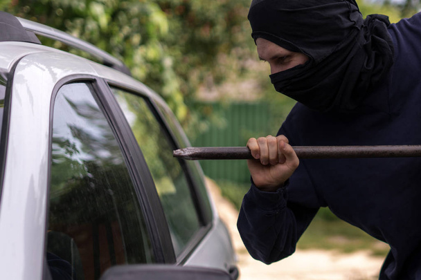 masked thief standing near the car and trying to smash the window with cowbar - Foto, Bild