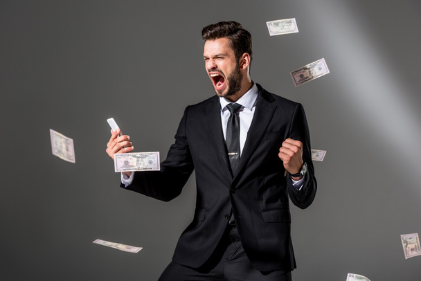 excited businessman with smartphone near falling dollar banknoteson on grey - Foto, Imagen