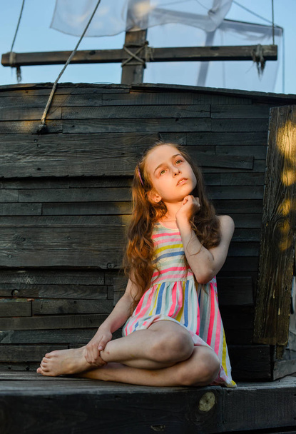Criança pequena pensativa com cabelos longos descansando no convés de madeira, olhando para o lado enquanto sonhava com algo
. - Foto, Imagem